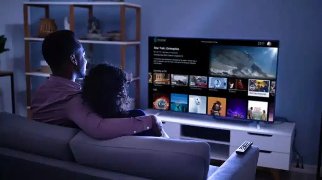 A couple sitting on a gray couch, viewed from behind, watching a large TV screen displaying a streaming service interface with multiple movie and show thumbnails.